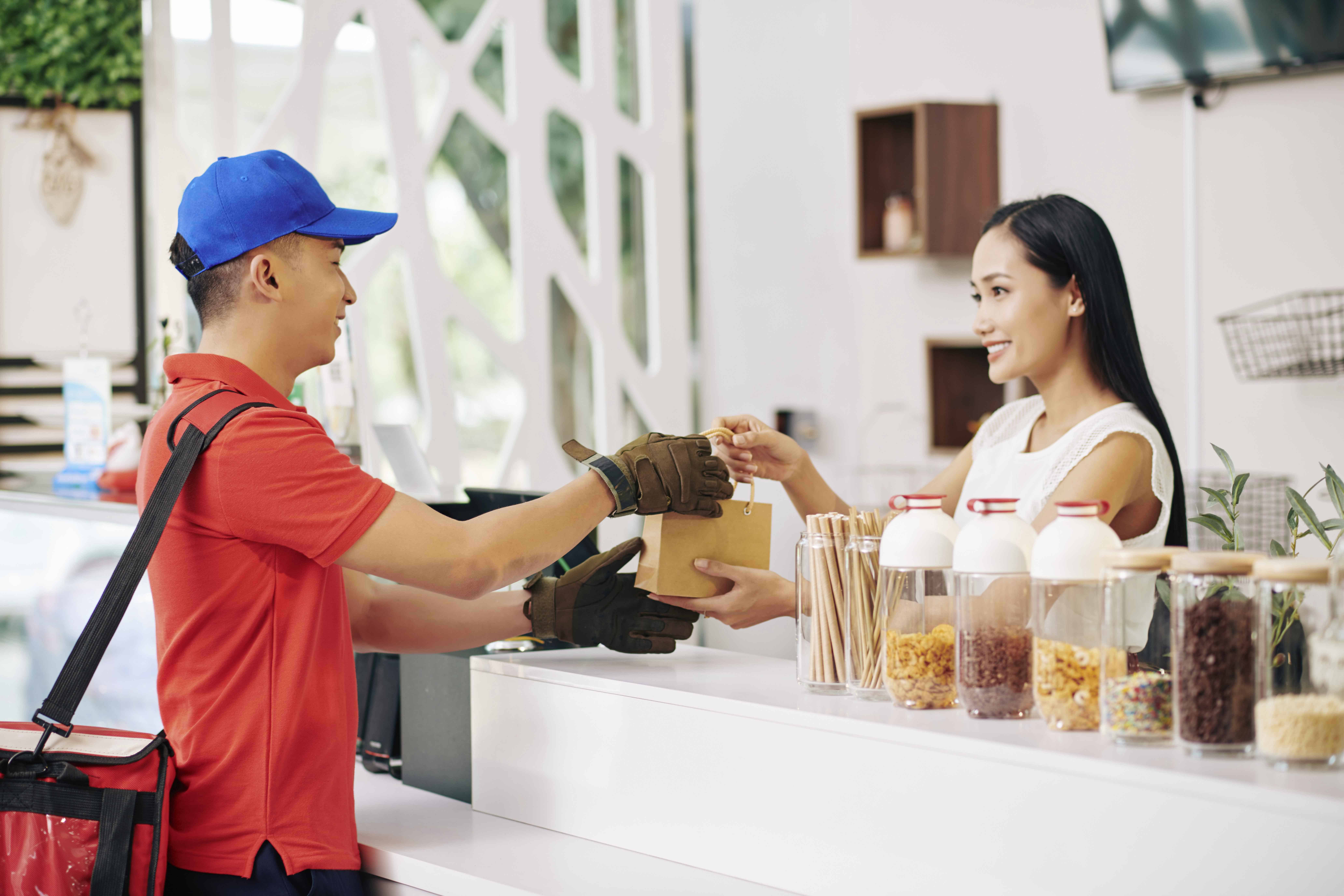 Smiling pretty small coffeeshop owner giving packed order to delivery man
